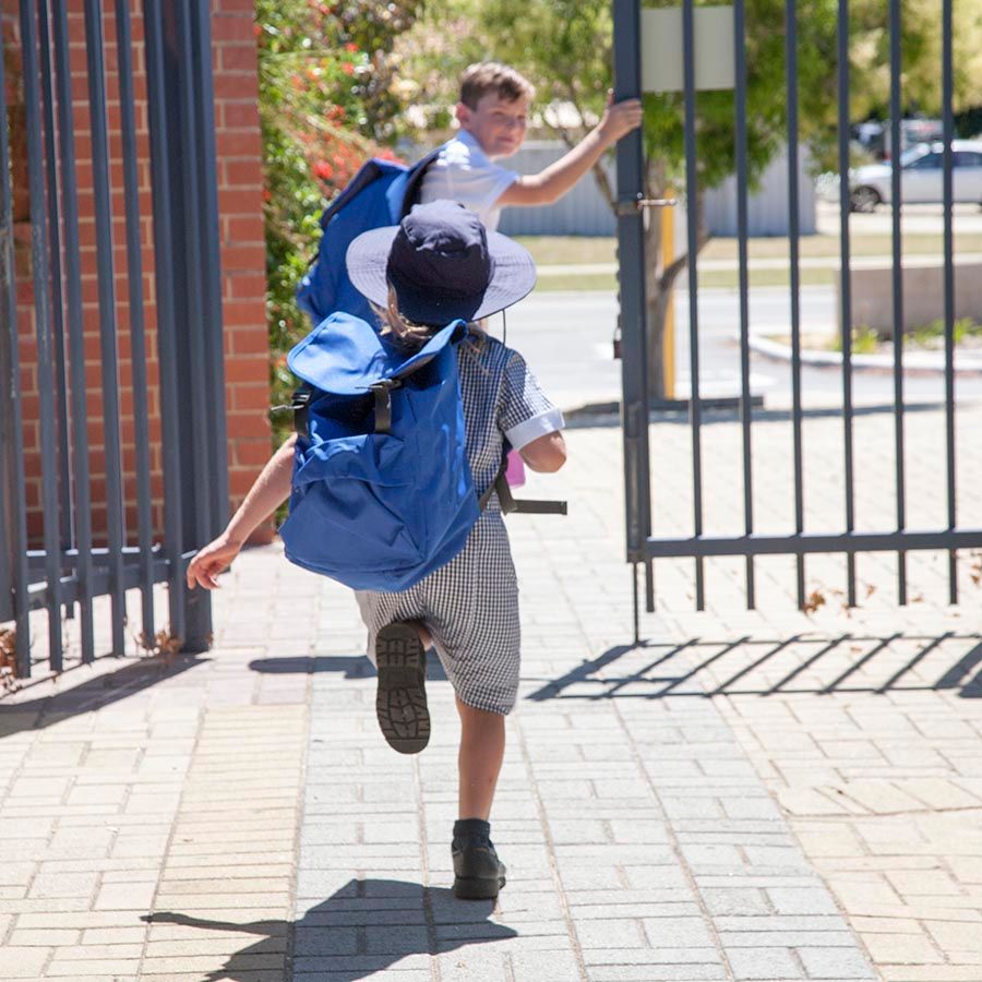 Two children are running out of tall metal school gates. Ahead is a boy in a white shirt with a backpack on, looking back as he holds onto the side of the gates, and in the mid-ground of the image is a child running with a school hat on, and with a blue backpack.